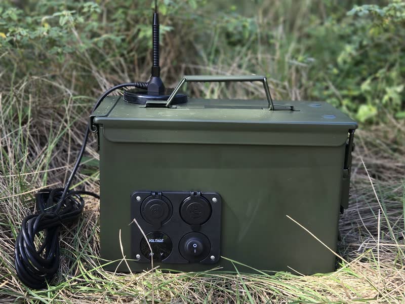 Closed olive-green ammo-can go-kit in tall grass, with an antenna on the lid and a front panel of power ports, a voltage readout and a switch.