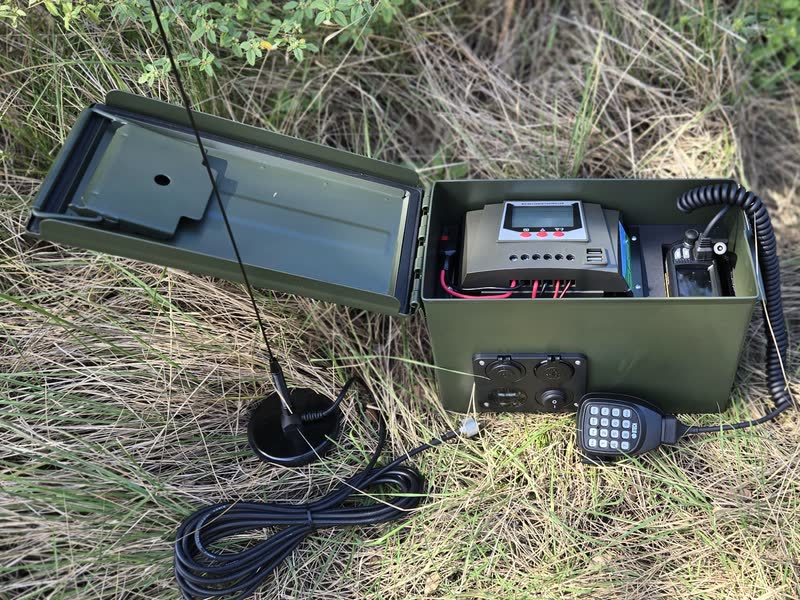 Ammo-can go-kit with the lid open, showing a radio and a small display unit mounted inside, power ports on the front, and a hand microphone and antenna on the grass beside it.