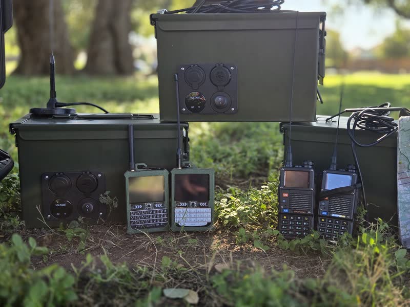 Three ammo-can go-kits stacked on the grass, with two small keyboard devices and two handheld radios in front of them.