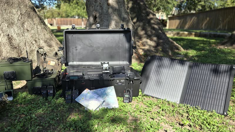 Radio kits laid out on the grass under oak trees: green ammo-can go-kits, an open black Pelican case with radio gear mounted inside, a folding solar panel, handheld radios and a paper map.