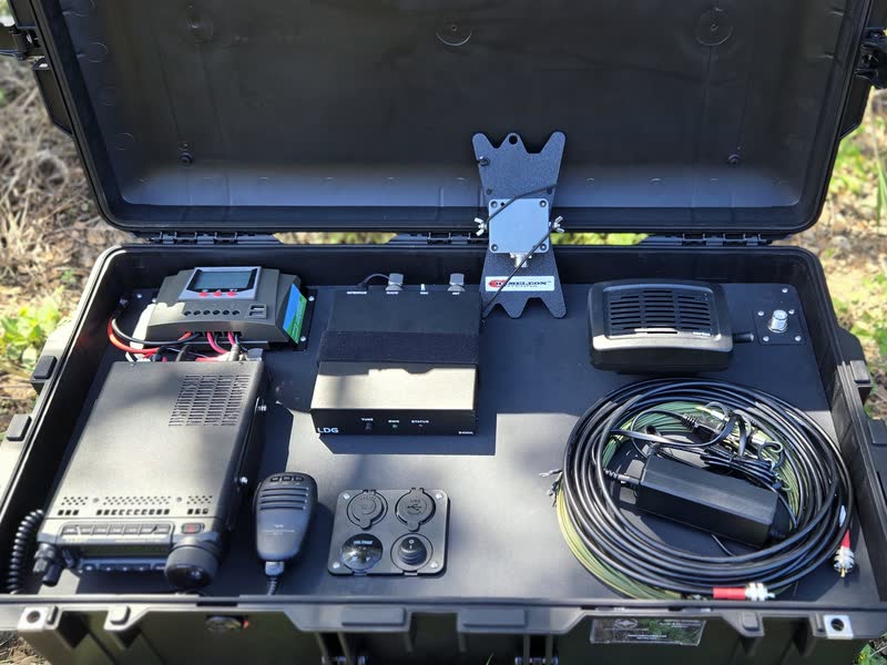 Inside the open Pelican case: a radio with a hand microphone, a small display unit, a speaker, power ports and a coil of antenna wire, all mounted on a black panel.