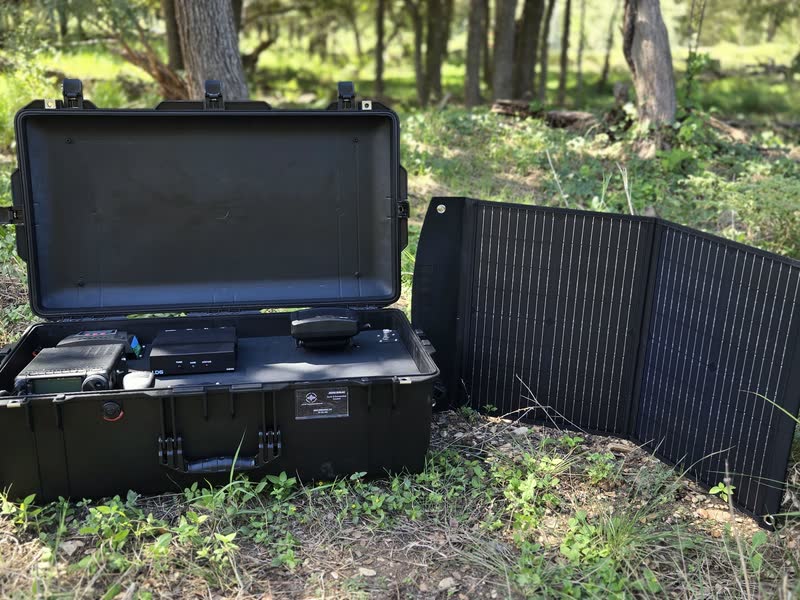 Black Pelican Air case radio kit open in a wooded field, with a folding solar panel standing beside it.